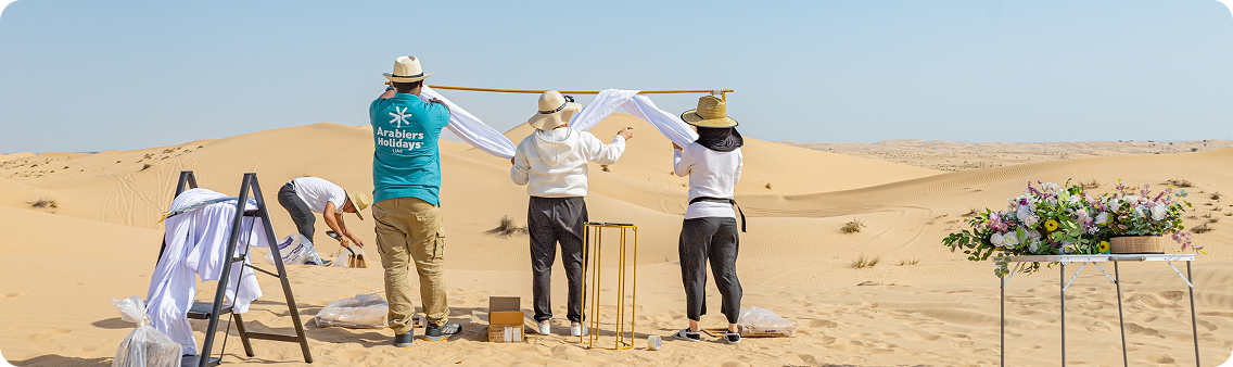 Professional team setting up private desert wedding venue in Dubai with floral arch installation on sand dunes